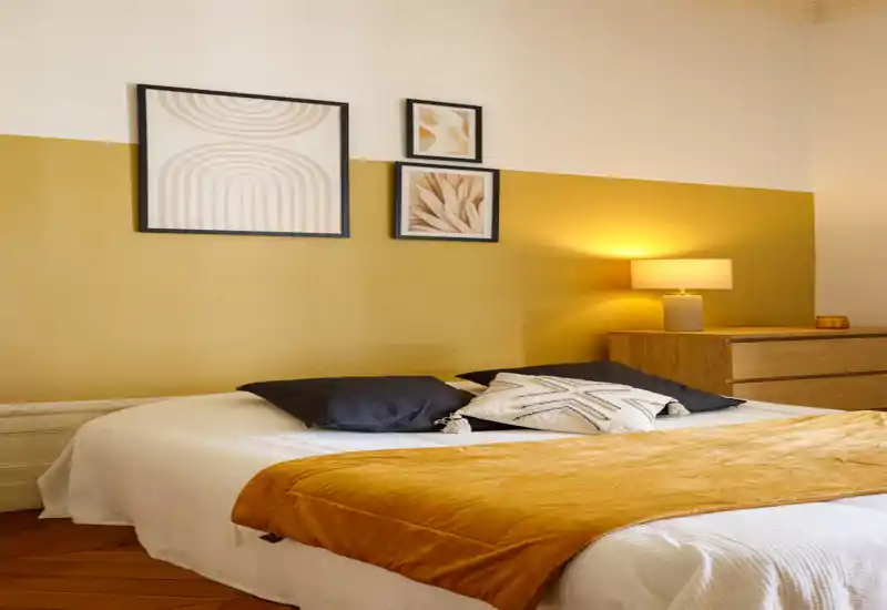 Bright, styled bedroom with a mustard accent wall, cozy throw and decorative cushions; warm bedside lamp and framed art create a welcoming atmosphere.