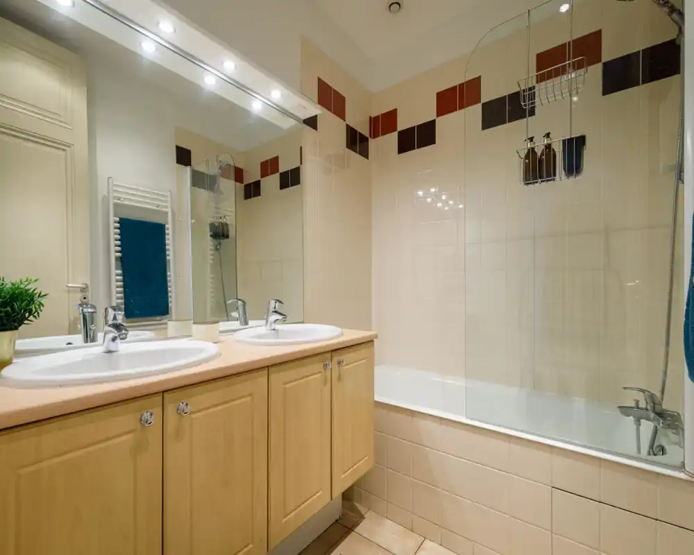 Bright, modern bathroom with a double sink vanity, large mirror and a bathtub with a glass screen. Neutral tiles and warm wood cabinetry create a clean, welcoming feel.
