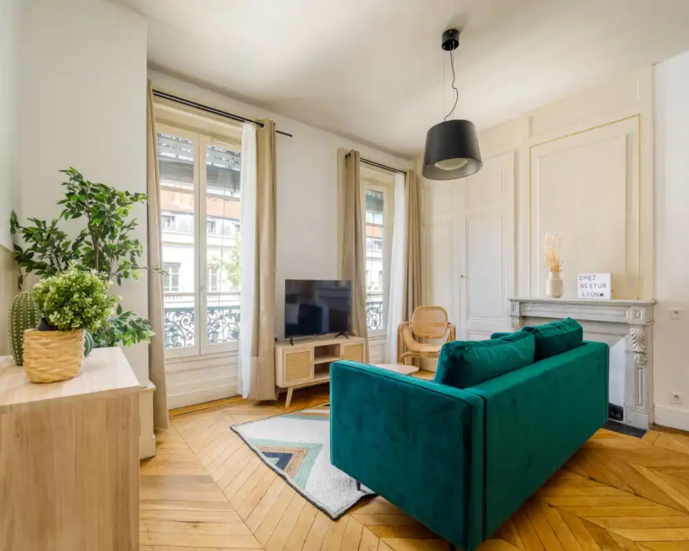 Bright, stylish living room with a deep emerald velvet sofa, natural light from tall windows, parquet floors and tasteful decor creating a welcoming lounge area.