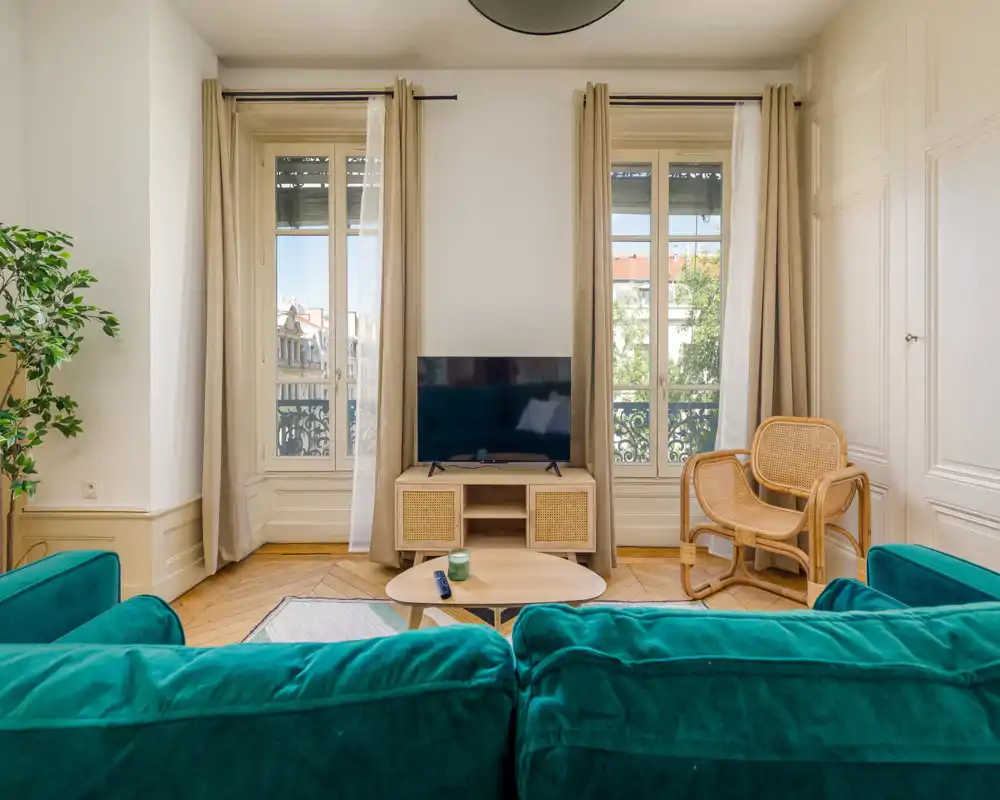 Sunlit living room with a teal sofa, a low wooden coffee table and TV console placed between two tall windows; warm parquet flooring and rattan chair add a natural, cozy touch.