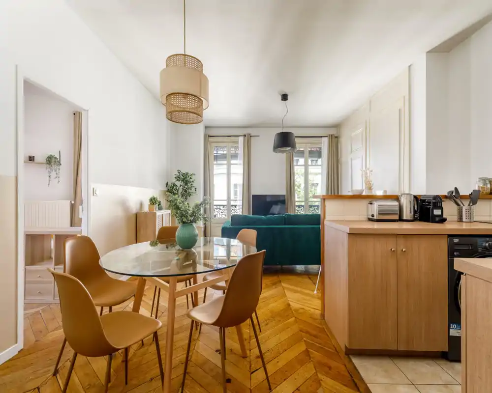 Spacious, sunlit living area with a modern teal sofa, large windows and a dining table — warm wooden floors and tasteful décor create a welcoming atmosphere perfect for rentals.