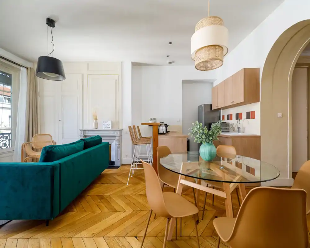 Bright, stylish open-plan living area with a teal velvet sofa, wooden herringbone floor and a glass dining table — modern Parisian-style living space ideal for rentals.