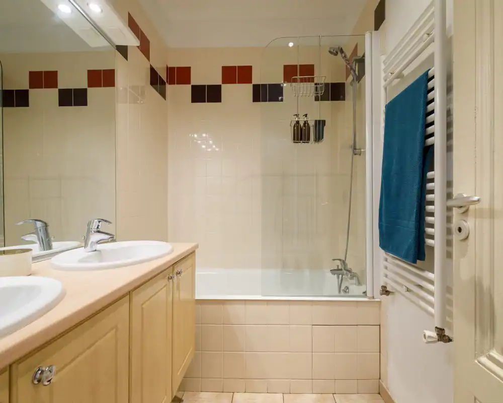 Bright, tidy bathroom with a bathtub and shower screen, dual sinks set into a light wood vanity and a heated towel rail with a blue towel.