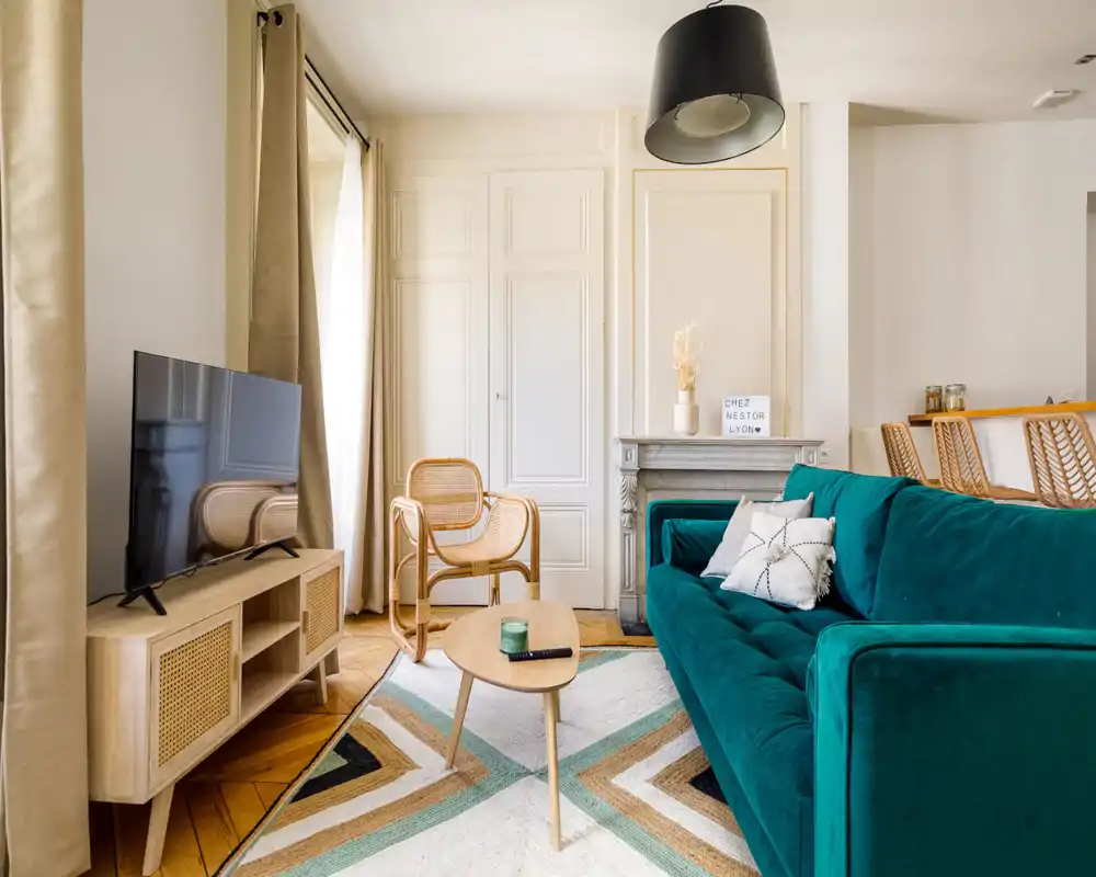 Bright, stylish living room with a deep teal sofa, natural rattan chairs, patterned rug and TV console — a cozy, well-lit space ideal for relaxing.