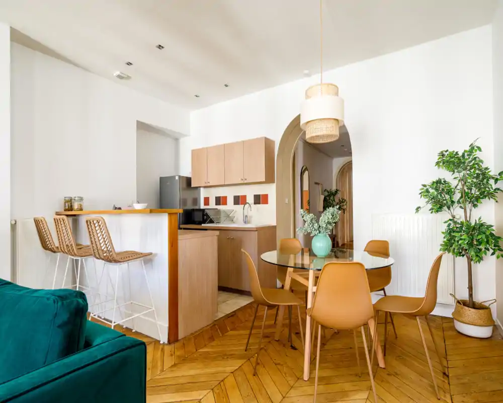 Bright, stylish open-plan living and dining area with wooden chevron floors, a glass dining table with modern chairs, a cozy teal sofa edge visible, and a compact kitchen with bar seating — ideal for social stays.