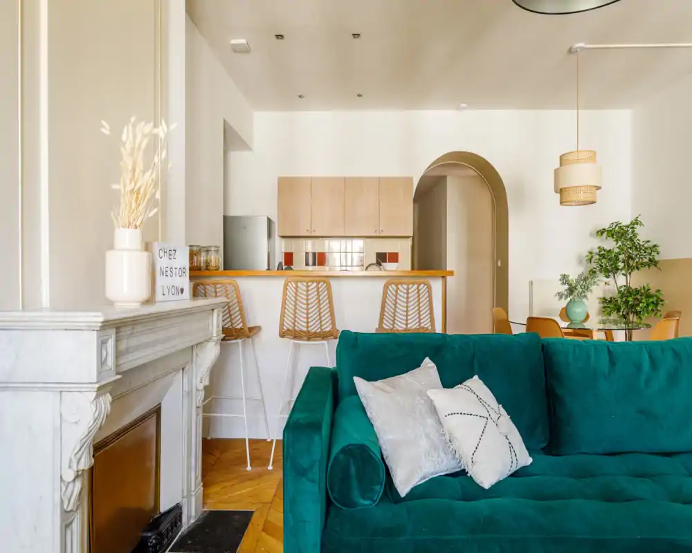 Stylish living area featuring a deep green velvet sofa, breakfast bar with rattan stools and an open sightline to a compact kitchen; warm wood floors and soft natural light create a welcoming atmosphere.