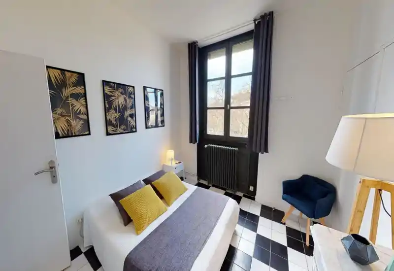 Bright, stylish bedroom with a double bed, decorative cushions and wall art. Natural light from a large window and tasteful black-and-white tiled floor create a modern, cozy atmosphere.