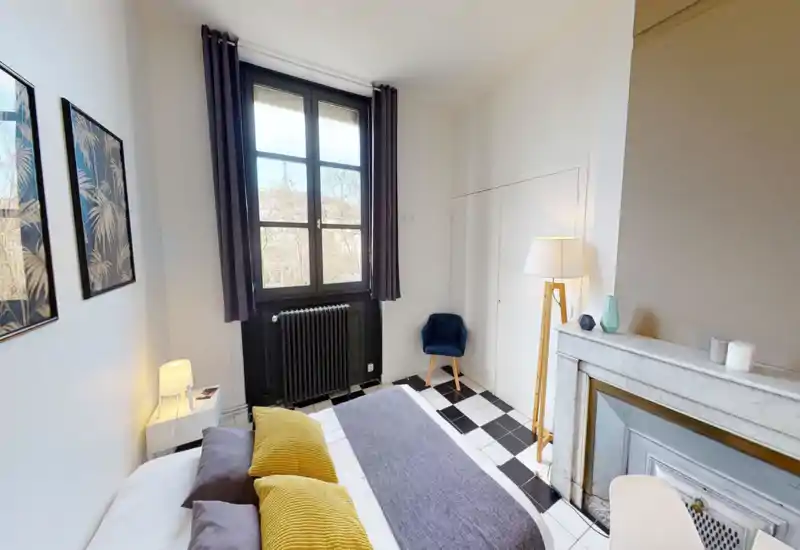 Bright, stylish bedroom with a large window, decorative pillows and a cozy atmosphere. The room features patterned floor tiles, a small armchair and a classic mantel adding character.