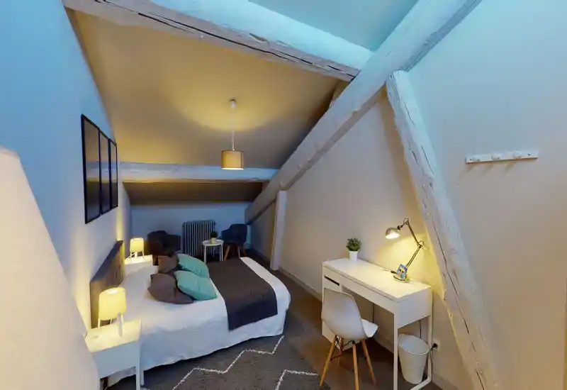 Accogliente camera mansardata con travi bianche a vista, letto matrimoniale con biancheria neutra e cuscini colorati, lampade da comodino e piccolo scrittoio — luminosa e ben curata, ideale per un annuncio.