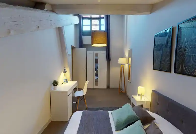 Ampia camera da letto in stile loft con travi a vista, letto matrimoniale confortevole con cuscini e copriletto, scrivania e armadio, illuminazione soffusa — luminosa e ben arredata per un soggiorno rilassante.