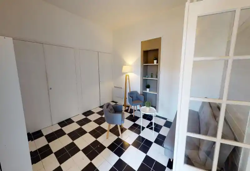 Bright, modern living area with two upholstered armchairs, a small round coffee table and a floor lamp on a stylish black-and-white tiled floor; shelving with plants and a sofa visible through glass doors complete the cozy sitting space.