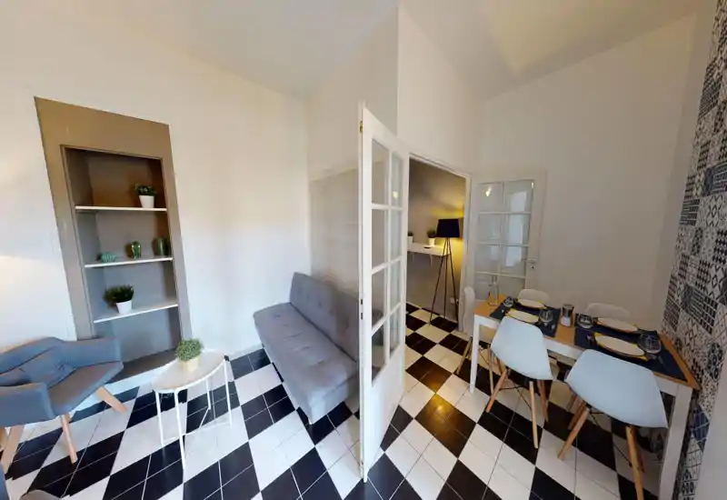 Bright, modern living/dining space with a grey sofa and small dining table set on a striking black-and-white tiled floor. The room features cozy accents — plants, a floor lamp and patterned wall — creating an inviting atmosphere for guests.