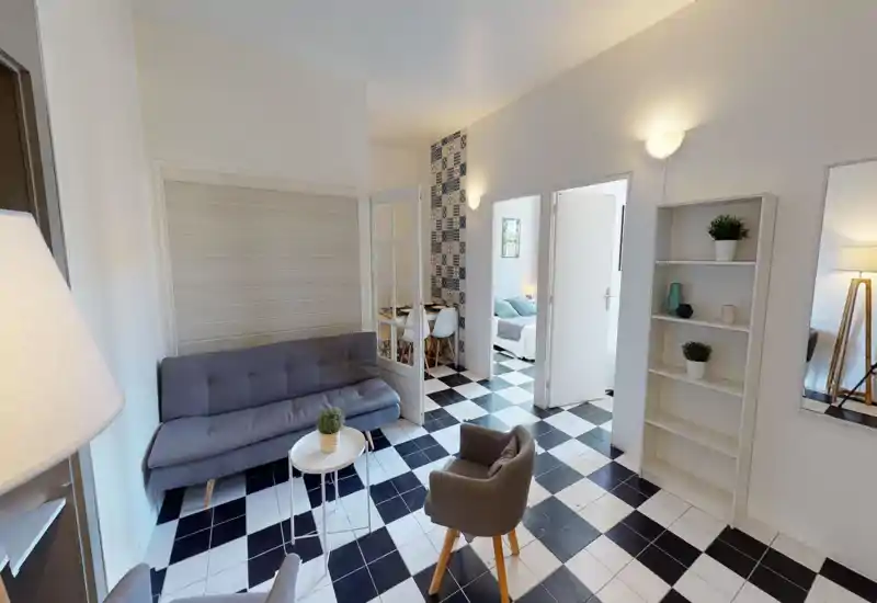 Bright, stylish living area with a gray sofa, armchair and checkerboard tile floor; tasteful decorative plants and shelving create a welcoming, well-staged space.
