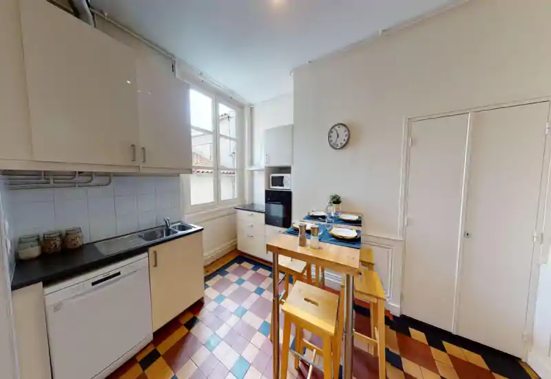 Bright, compact kitchen with modern cabinets, a sink and dishwasher on the left, and a small high dining table set for four. Large window provides abundant natural light and colorful tiled flooring adds character.