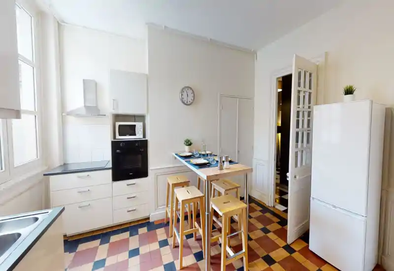 Bright, modern kitchen with white cabinets, built-in oven and microwave, tall fridge and a wooden breakfast bar with stools — well-lit by a large window and neatly staged for guests.