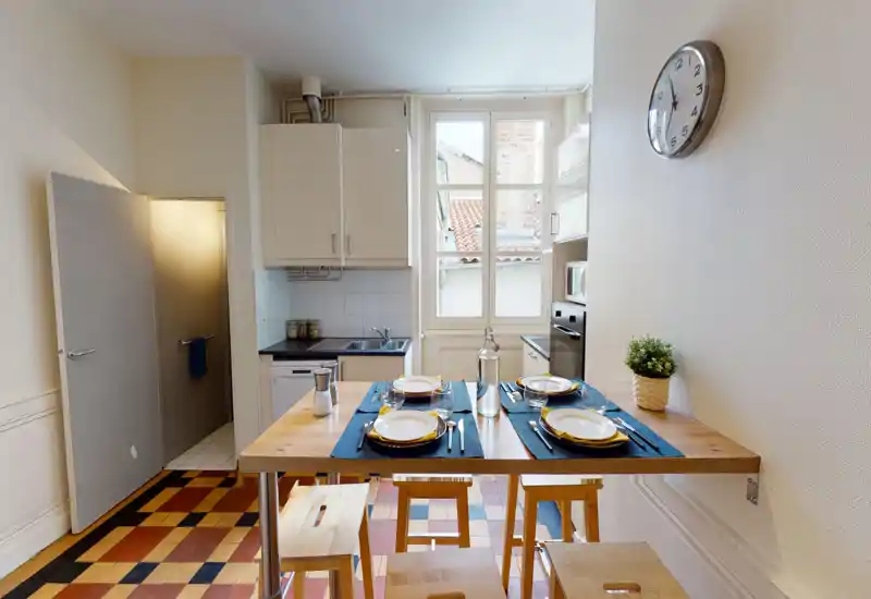 Bright, compact kitchen with a wooden breakfast table set for four, white cabinets, built-in appliances and a large window that brings in natural light.