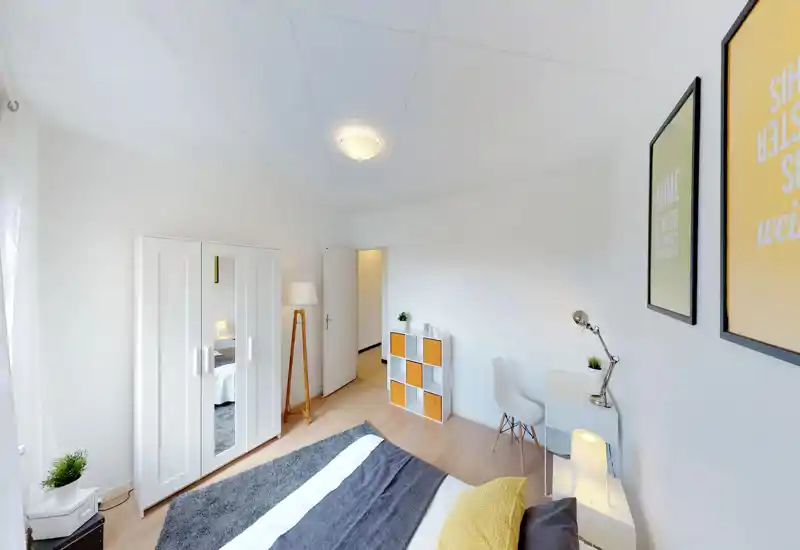 Camera da letto luminosa e moderna con letto ben rifatto, morbido tappeto e accenti gialli coordinati; comprende armadio, scrivania e luce naturale dalla finestra.