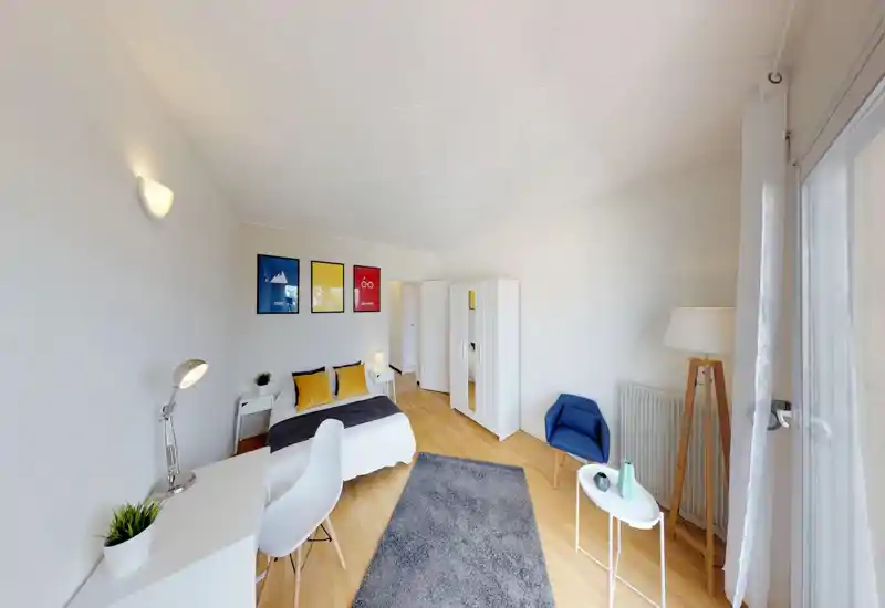 Camera da letto ampia e luminosa con letto matrimoniale, cuscini decorativi, scrivania e armadio; grande finestra con molta luce naturale e angolo salotto accogliente con poltrona e lampada.
