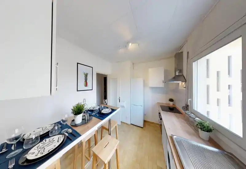 Cucina moderna e luminosa con piano in legno, piano cottura, frigorifero e un bancone alto apparecchiato per la colazione. La grande finestra illumina l'ambiente e le piante rendono l'aria familiare.