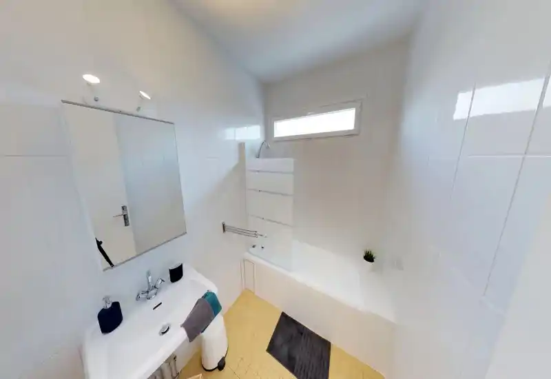Salle de bains lumineuse et moderne avec murs carrelés blancs, baignoire avec pare-douche en verre, miroir mural et lavabo avec accessoires. Style épuré et lumière naturelle provenant d'une fenêtre haute.