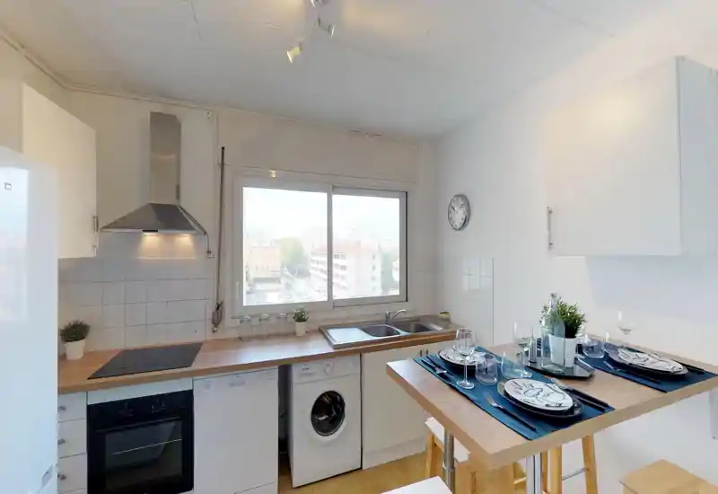 Cuisine lumineuse et moderne avec électroménagers intégrés, évier double sous une grande fenêtre et bar en bois dressé pour quatre. Placards blancs et décoration sobre pour un espace fonctionnel adapté aux locations courtes durées.