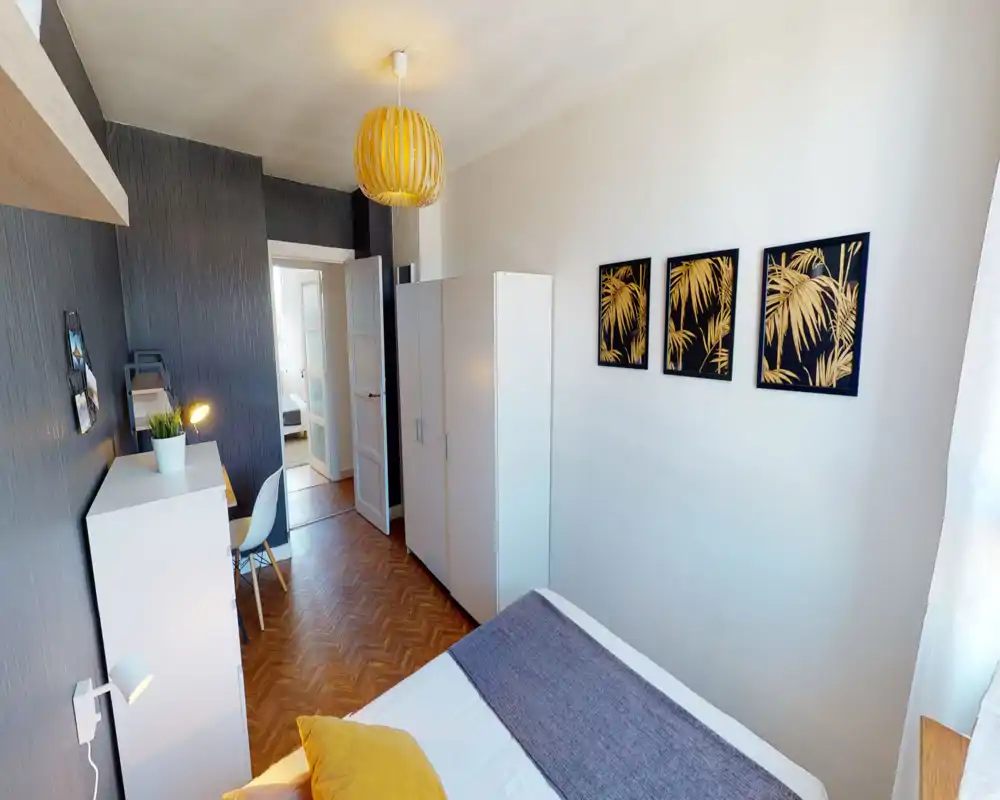 Chambre lumineuse et soignée avec un lit double visible, lampes de chevet et armoire. Parquet chaleureux, coin bureau et tableaux décoratifs apportent une atmosphère cosy.
