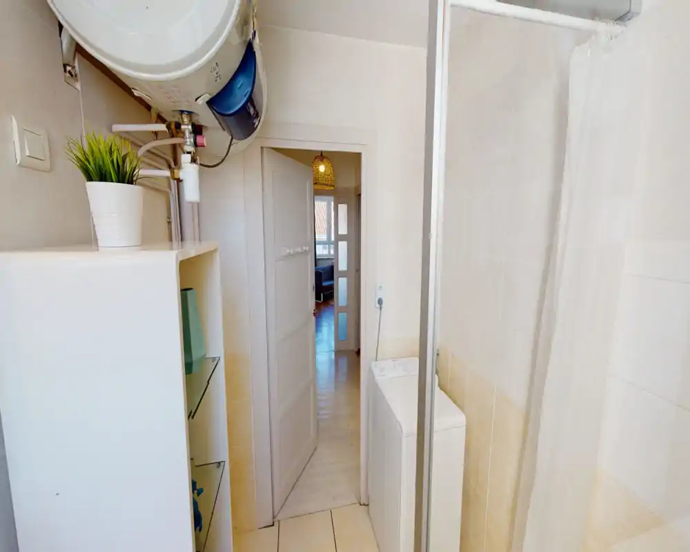 Salle de bain compacte aux carreaux clairs, douche avec rideau et machine à laver ; espace lumineux et propre adapté à un petit appartement.
