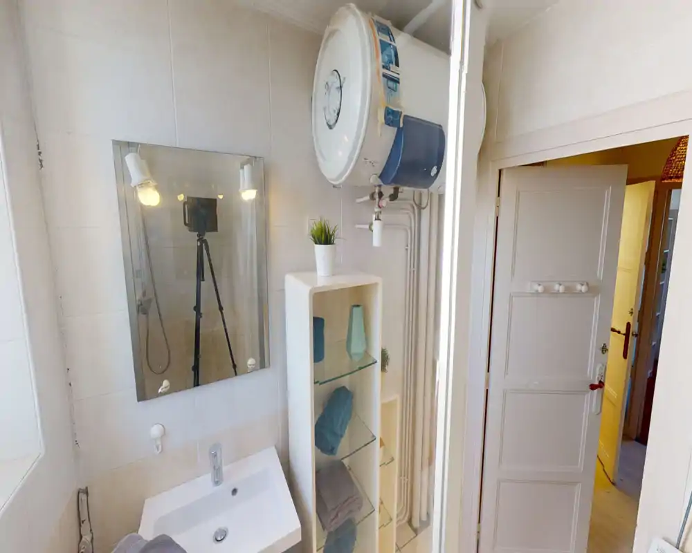Salle de bains compacte et lumineuse avec lavabo, miroir et étagères ouvertes contenant serviettes et objets déco ; chauffe-eau visible et carrelage propre.