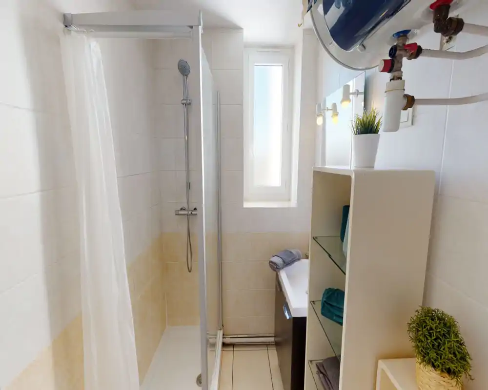 Salle de bains moderne et lumineuse avec une douche carrelée, pare-douche en verre, petit lavabo et étagères ouvertes avec serviettes et plantes.