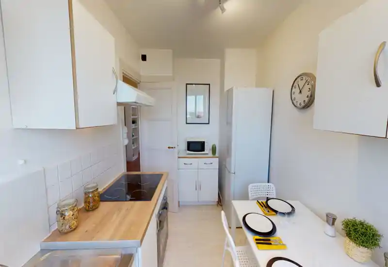 Bright, compact kitchen with white cabinets, electric cooktop, fridge and a small dining table set for two. Clean, modern styling and practical work surfaces ideal for a small apartment.