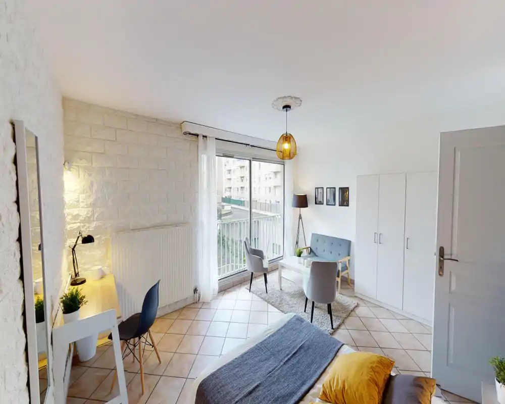 Bright, well-staged studio bedroom with a comfortable bed in the foreground, a small seating area by the window and a workspace. Large sliding window provides abundant natural light and a pleasant view onto the neighboring buildings.