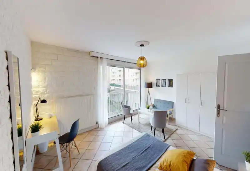 Bright, well-staged studio bedroom with a comfortable bed in the foreground, a small seating area by the window and a workspace. Large sliding window provides abundant natural light and a pleasant view onto the neighboring buildings.