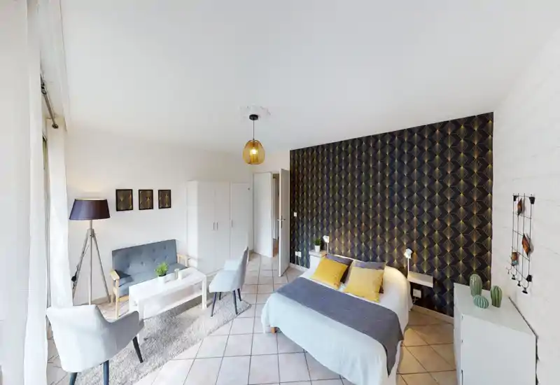 Stylish studio bedroom with a comfortable double bed, patterned accent wall and a cozy seating area by a bright window — ideal for short-term rentals.