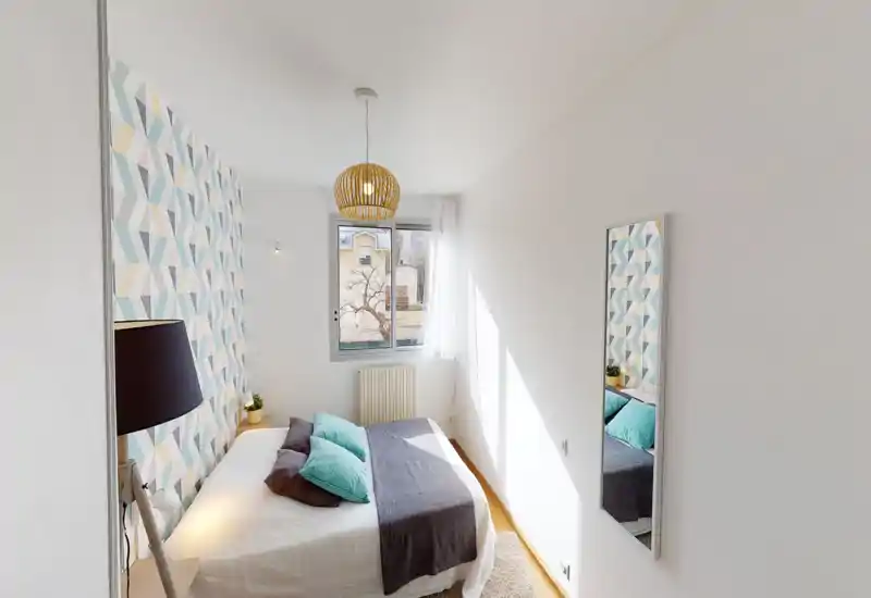 Bright, modern bedroom with patterned accent wall, double bed dressed in grey and teal textiles, natural light from a window and a full-length mirror reflecting the décor.
