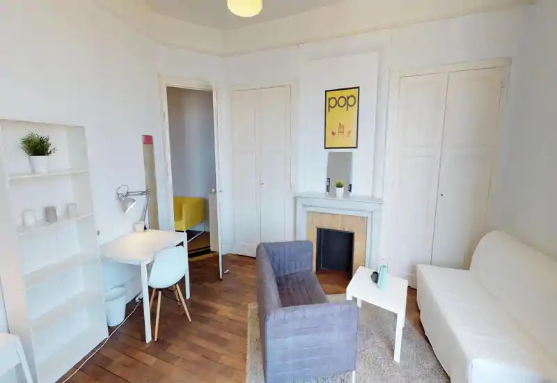 Bright and cozy living room with a small sofa, armchair, coffee table and a desk area; white walls and warm wooden floors create a welcoming space.