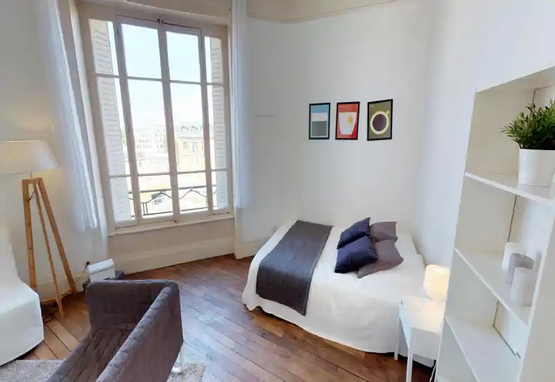 Bright, cozy bedroom with a double bed dressed in white linen, decorative pillows and a dark runner; large window floods the room with natural light and wooden floors add warmth.