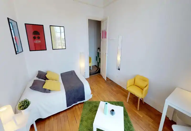 Chambre lumineuse et bien aménagée avec un lit double, coussins colorés, tapis vert et un fauteuil confortable; parquet et affiches murales apportent une ambiance accueillante.