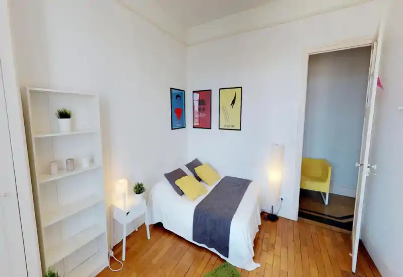 Chambre lumineuse et soignée avec un lit double en linge blanc, jeté gris et coussins jaunes, étagère minimaliste et affiches encadrées — idéale pour une annonce chaleureuse.