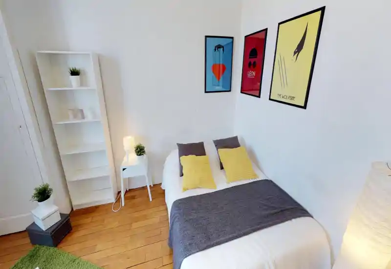 Chambre lumineuse et soignée avec un lit double en linge blanc, plaid gris et coussins jaunes ; des affiches murales et une table de chevet avec lampe apportent une ambiance moderne et chaleureuse.
