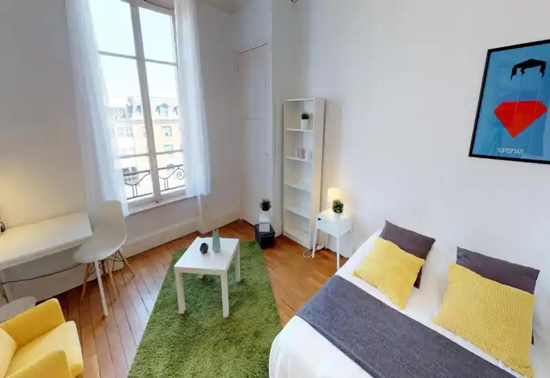 Chambre lumineuse et moderne avec un lit double en linge blanc et coussins jaunes, un tapis vert, une petite table basse et un bureau près d'une grande fenêtre apportant beaucoup de lumière naturelle.