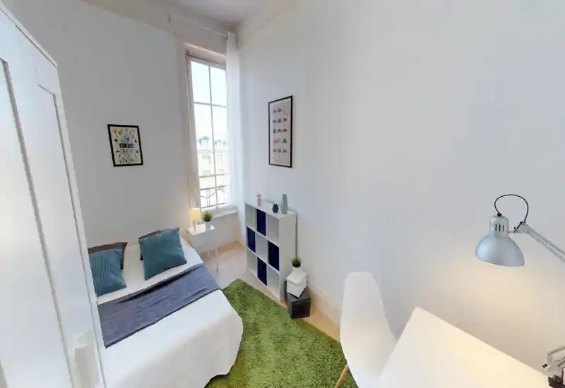 Bright, compact bedroom with a double bed dressed in white linen, blue cushions and a green rug; natural light from a tall window and a small desk create a neat, Scandinavian-style feel.
