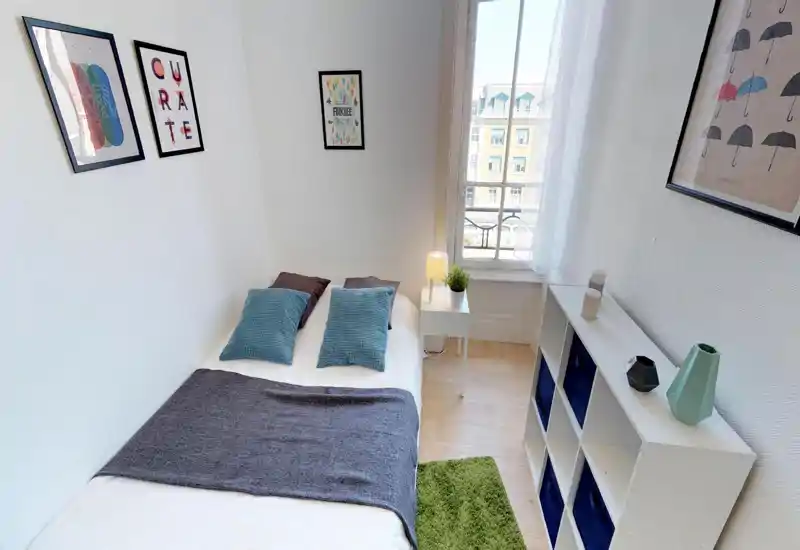 Bright, compact bedroom with a neatly made double bed, decorative cushions, a bedside table with lamp and plant, and a shelving unit for storage. Large window provides good natural light and a pleasant street view.