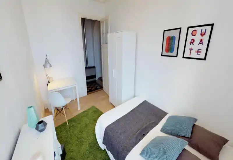 Bright, compact bedroom with a comfortable double bed, textured cushions, green rug and a small desk. White walls, wardrobe and framed prints create a fresh, minimalist look ideal for short-term rentals.