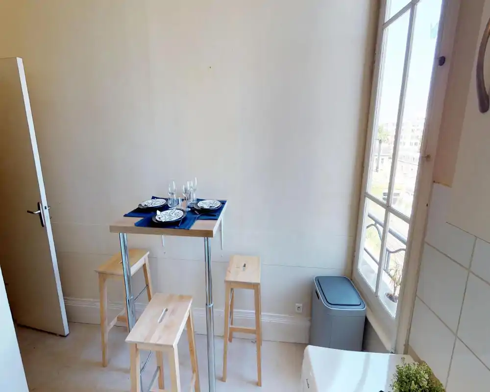 Compact dining corner with a high wooden table and three stools set with plates and glasses, bright natural light from a tall window and a minimal, modern feel.