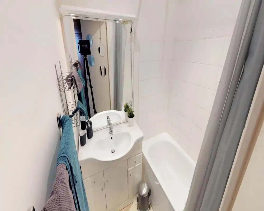Compact, well-lit bathroom with a sink cabinet, mirror, bathtub with shower curtain, and towel racks. Clean white tiles and neutral decor create a fresh, functional space ideal for short stays.
