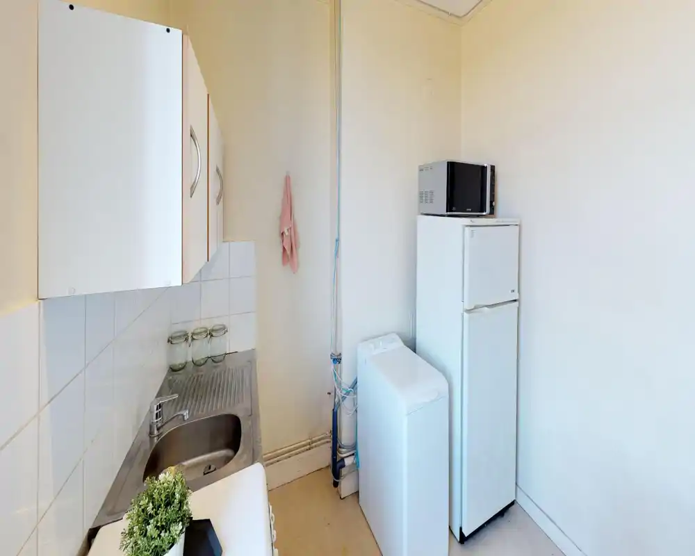 Compact kitchen corner with a stainless sink, wall cabinets, refrigerator, microwave and a top-loading washing machine; bright, functional layout suitable for a small apartment.