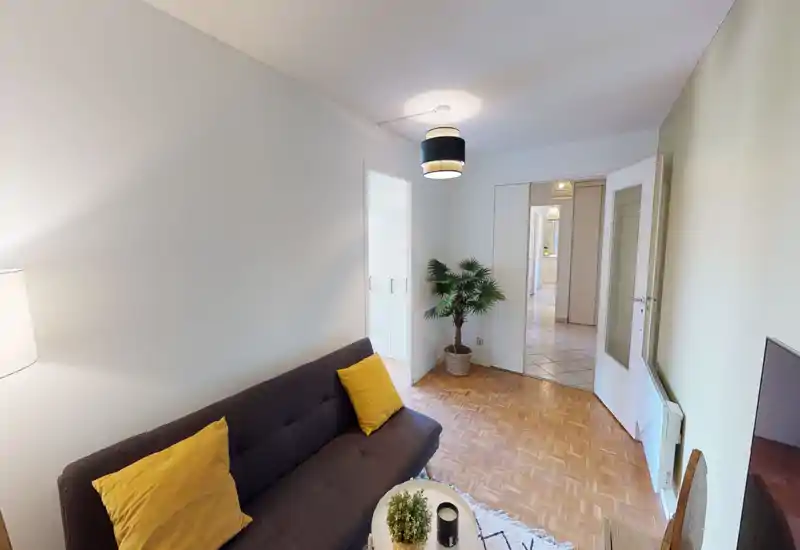Bright, neatly staged living room with a compact dark sofa, yellow accent cushions, a small coffee table and a decorative potted plant — ideal for a cosy rental lounge.