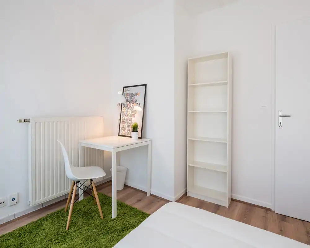 Rincón de dormitorio luminoso y minimalista con la cama blanca en primer plano, un pequeño escritorio con lámpara, una estantería sencilla y una alfombra verde que aporta color.