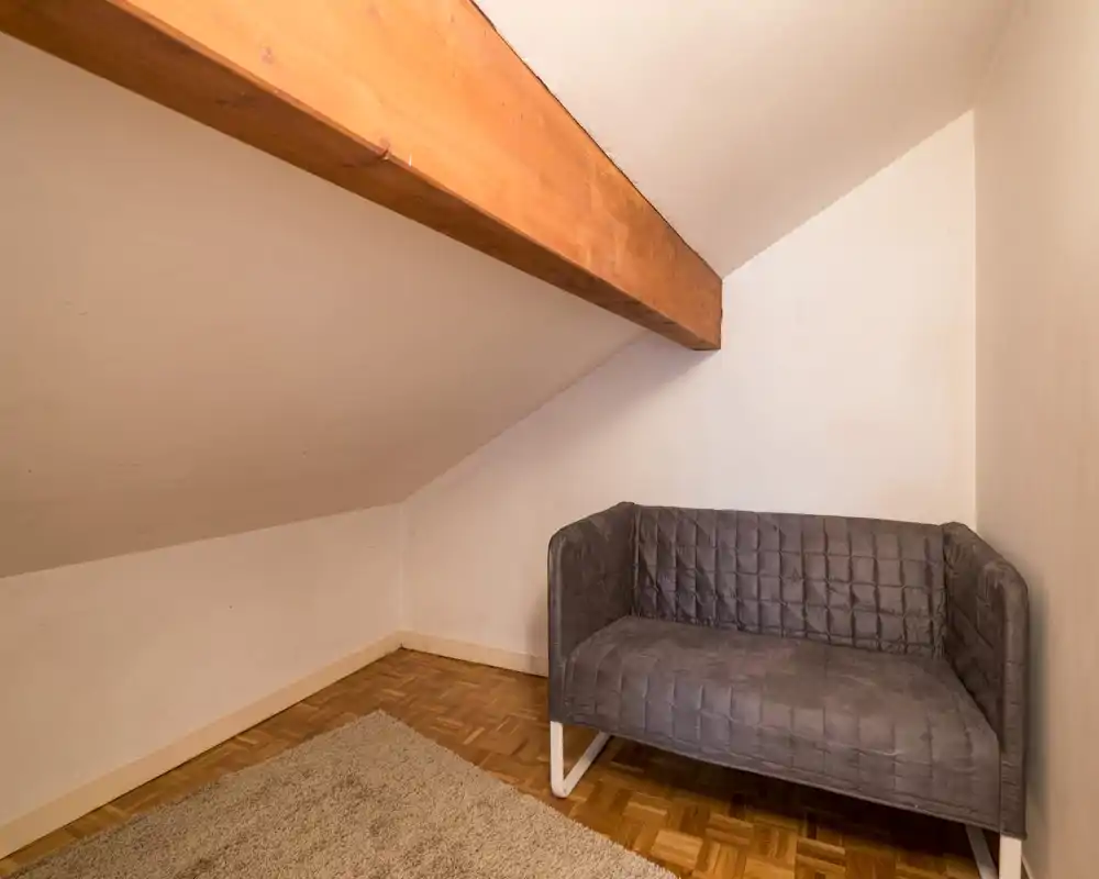 Compact attic living area with a small grey sofa, exposed wooden beam and parquet flooring creating a cozy corner ideal for reading or relaxing.