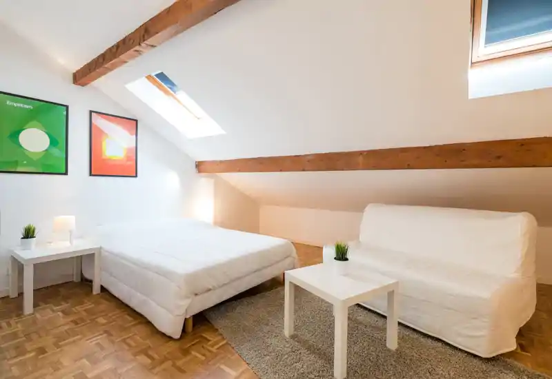 Chambre mansardée lumineuse avec poutres apparentes, lit double bien fait et petit canapé. Les fenêtres de toit et la décoration minimaliste offrent une ambiance aérée et chaleureuse, idéale pour des courts séjours.